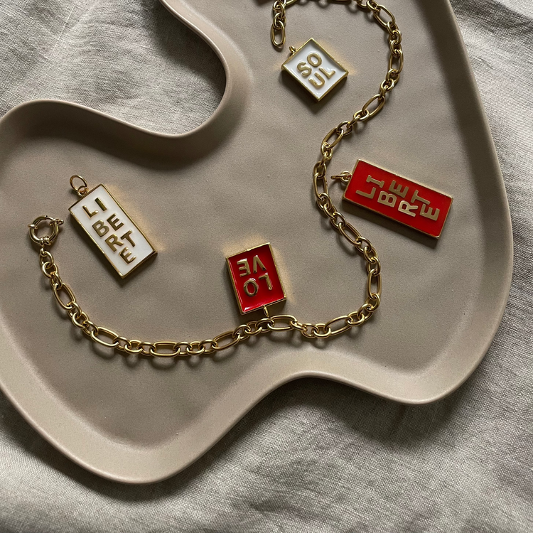 Mehrere Anhänger auf einem Tablett. Die Anhänger haben einen golderen Rahmen und farbige füllungen. In der Füllung aus Kunstharz sind vergoldete Buchstaben eingelassen, die Statements wie LOVE und Liberté darstellen